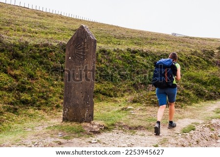 Pilgrim walking next to the Stone Post with Scallop Shell Symbol of the Pilgrimage in the Pilgrim Trail of the Way of St James or Camino de Santiago. French Pyrenees