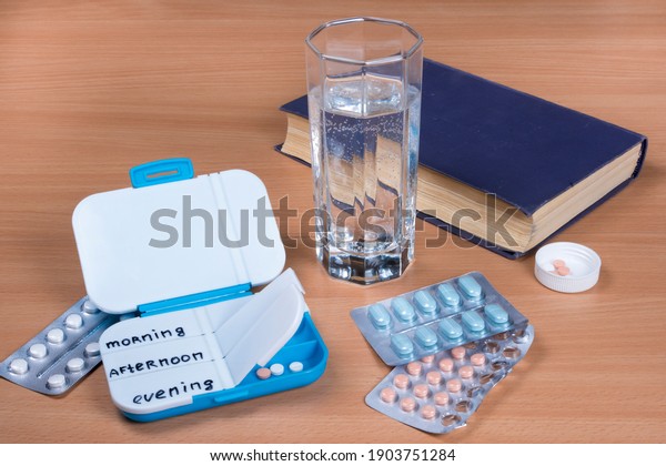 Pill box daily take medicine with pills, glass of water\
and book. Medication in hospital on wooden table. Drugs use for\
treatment. 