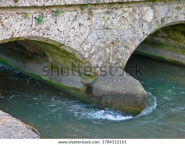 The\
pillar of a small bridge resists the small river and divides the\
water that flows under the arches of the\
bridge.