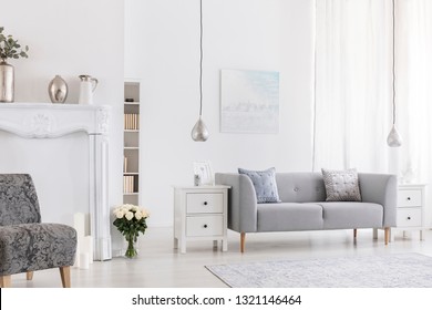 Pillows on grey settee in white living room interior with flowers near patterned armchair. Real photo