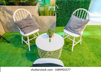 A pillows on white wooden chairs and a artificial tree in pot on a white table on artificial grass with nature hedge.