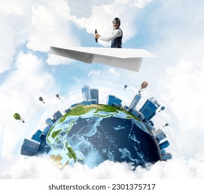 Pilot in leather helmet and goggles driving paper plane in cloudy blue sky. Funny man flying in small paper airplane over earth globe. Spherical view of modern city with high skyscrapers. - Powered by Shutterstock