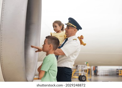 Pilot In Uniform Showing Parts Of Airplane To Two Little Kids Who Came To Excursion At The Aircraft Hangar. Aircraft, Excursion, Childhood Concept