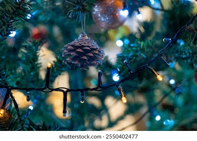 A pine cone hangs delicately from a string of lights on a Christmas tree, surrounded by colorful ornaments and warm glowing lights in a festive atmosphere. - Powered by Shutterstock