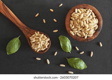 Pine nuts overhead shot with a close up top view of many shelled pine nuts in a wooden spoon and in a bowl on a dark grey background.