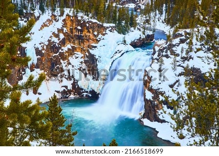 Pine trees frame snowy cliffs and stunning blue waterfalls in Yellowstone