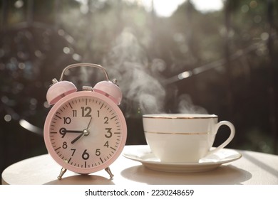 Pink alarm clock and cup with hot drink on white table in morning