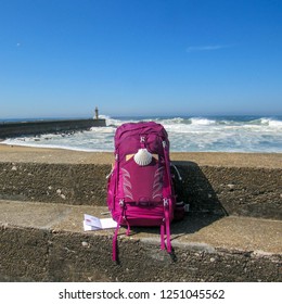 Pink backpack with scallop shell pilgrim symbol at concrete wall with ocean with huge white waves and blue sky on background. Pilgrimage route Camino de Santiago or Way of Saint James, Porto, Portugal