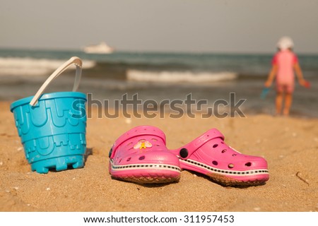 Pink beach crocs and blue sand toys on sandy beach.Beach flip flops in the foreground and blurred sea in the background. 