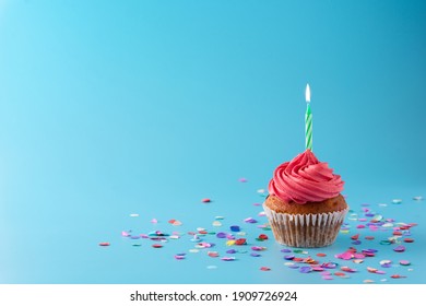 Pink birthday cupcake with green candle and colorful confetti on blue background. Copy space