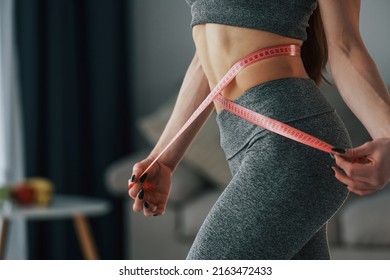 Pink Colored Measuring Tape Around The Waist. Young Woman With Slim Body Type And In Yoga Clothes Is At Home.