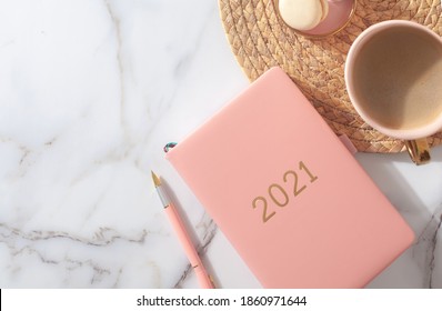 Pink coral colored diary for the year 2021, pen, coffee latte, macaron cookie and straw woven placemat on white marble background. New year planning concept. Minimalistic workstation. Copy space.