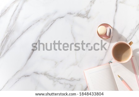 Pink coral colored diary for the year 2021, pen, coffee latte, macaron cookie and straw woven placemat on white marble background. New year planning concept. Minimalistic workstation. Copy space.