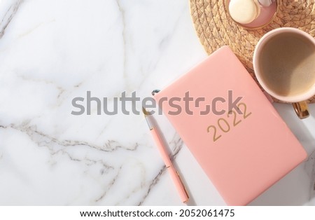 Pink coral colored diary for the year 2022, pen, coffee latte, macaron cookie and straw woven placemat on white marble background. New year planning concept. Minimalistic workstation. Copy space.