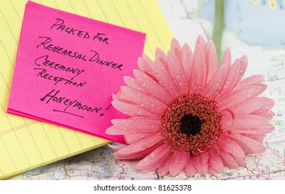 Pink daisy on map with note, "Packed for rehearsal dinner, ceremony, reception, and honeymoon" on top of map and notepad.