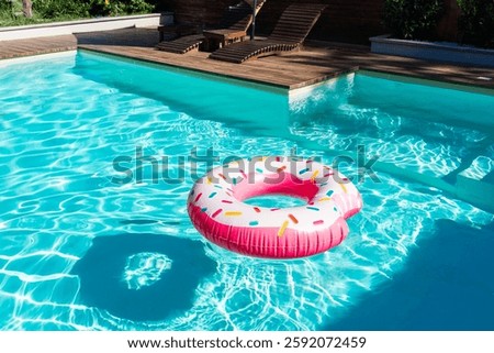 Pink donut-shaped inflatable float in turquoise swimming pool with wooden deck and sunny reflections