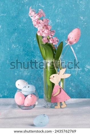 Pink Easter decorations decorative eggs, cute wooden bunny, jacinth flower in glass. Composition on white table with turquoise backdrop. Easter card. Vertical format.