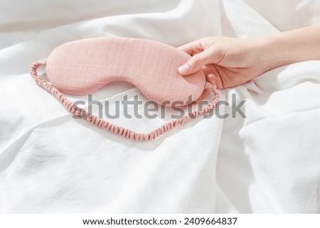 Pink eye mask for sleep on bed background, minimal lifestyle aesthetic scene, copy space. Top view woman hand holding sleeping mask on white bed linen, morning daylight. Comfort rest, healthy trend