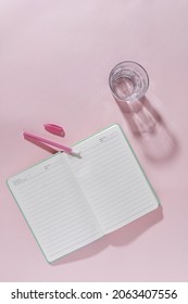 Pink Feminine Office Workspace Still Life With Open Journal Or Notebook, Pink Pen And A Glass Of Fresh Healthy Water In A Top Down Flat Lay View With Copy Space