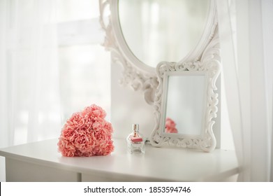 Pink flowers on the dressing table in the white bedroom. Flowers and perfumes in a bright interior. White boudoir table close up and copy space.