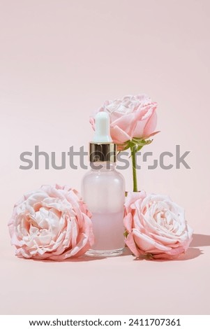Pink glass cosmetic bottle with a dropper on a pink background with rose flowers in bloom. Natural cosmetics concept, natural essential oil and skin care products.