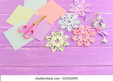 Pink, Green, Blue And Purple Paper Snowflakes. Paper Snowflake Set, Colored Paper Sheets And Scraps, Scissors On Wooden Background. Kids Winter Crafts