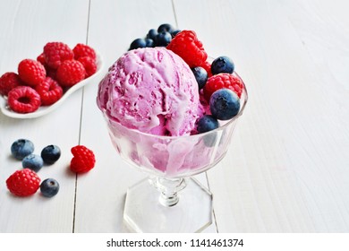 Pink ice cream raspberry and blueberry flavor topped with fresh berries in glass cup over white wooden table