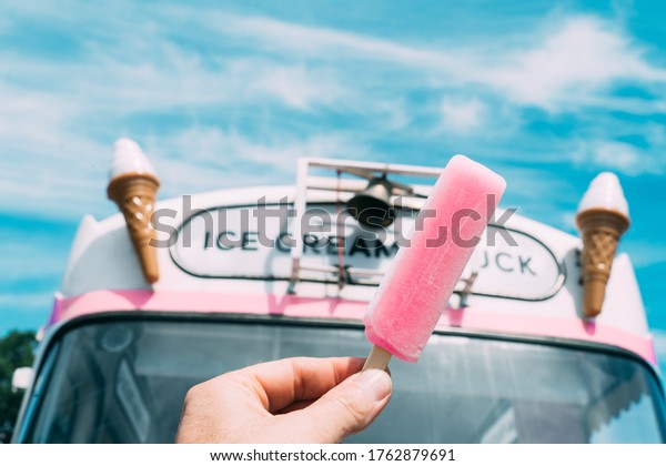 Pink ice cream with a pink ice
cream truck or van in the background. Hand holding ice
cream.