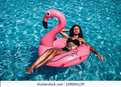 Pink inflatable ring shaped as flamingo and amazing African-American woman having suntan in the pool with her eyes closed; to view shot.