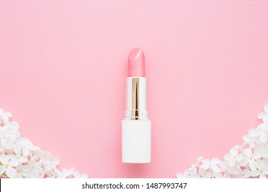 Pink lipstick in white tube with fresh lilac blossoms on light pastel background. Decorative cosmetic. Closeup.