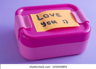 Pink Lunch Box With Orange Sticker On Purple Background. A Love You Message. Lunch From A Loving Wife Or Mother With A Note. Bright Colors And Care Concept. Idea For Food From Home Packaging. 