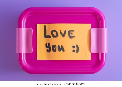 Pink Lunch Box With Orange Sticker On Purple Background. A Love Message With A Heart. Lunch At Work From A Loving Wife With A Note. Bright Colors And Care Concept. Idea For Food From Home Packaging.