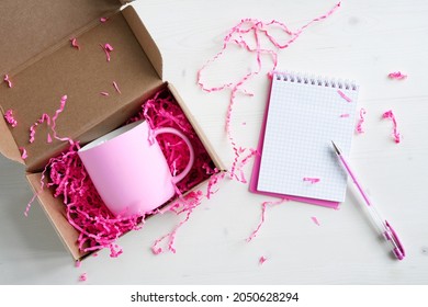 Pink Notebook, Pen, Mug And Box With Pink Crinkle Cut Paper Shred Filler On A White Table. List Of Gifts For The Holidays. Unpacking Gifts.