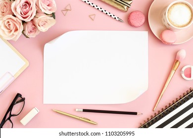 Pink office table with cup of coffee, flowers, macarons, pen, pencil, make up brush, notebook and empty paper. Woman's accessories, morning coffee, writing concept. Top view, flat lay, space for text.