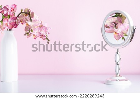 Pink orchids in vase reflected in mirror. Cosmetic dressing table. Light bathroom decor, mirror, pink orchids flowers on pink shelf. Elegant decor bathroom interior.