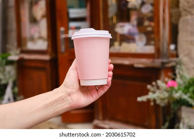 Pink Paper Cup With Coffee In A Woman's Hand. It's Time To Drink Coffee In The City. Disposable Paper Cup Close-up On The Background Of The City. Empty Space For Text, Layout. Coffee To Go Concept