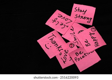 Pink paper notes with handwritten positive messages like "Smile," "Thanks," "I Love You," and "Be Happy," displayed on a black background, offering empty space for text. - Powered by Shutterstock