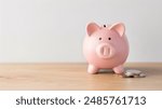 Pink piggy bank on wooden table, overflowing coins, clean white wall background. A pink piggy bank overflowing with coins is placed on a wooden table, against a white wall background.