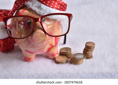 Pink Piggy Bank With Santa Claus Hat And Glasses. With Coins Around. In The White Snow. Christmas Savings Concept.