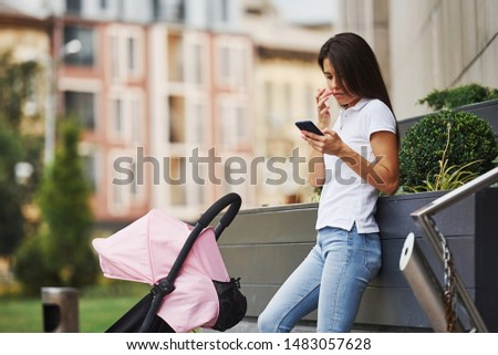 With pink pram. Using phone. Pretty young woman have weekend in the city at daytime.