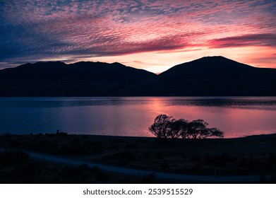 Pink and purple sunset sky over a calm lake with silhouetted hills and a tree. - Powered by Shutterstock