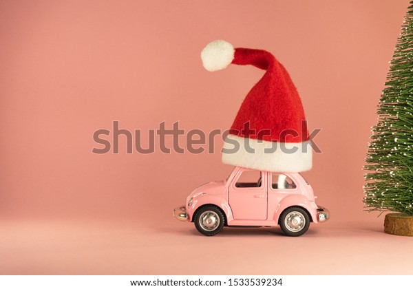 Pink retro toy model car with small\
red Christmas Santa Claus hat and little Xmas tree on pastel pink\
background. Vilnius, Lithuania, September 09,\
2019