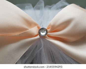 Pink satin bow of peach color with one small white round rhinestone in the center  on a white tulle fabric gathered in folds (macro, full face, texture).