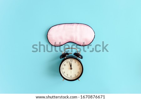 Pink sleep mask for eyes and black alarm clock on blue background It 's midnight. Top view Copy space. Concept eye protection from light for good sleep and melatonin production.
