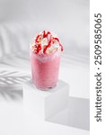 Pink strawberry milkshake topped with whipped cream and red syrup is standing on white cube on white background with shadows