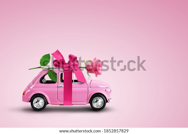 Pink toy car delivering pink rose flower with\
ribbon and bowon pink background. Valentine day, flowers delivery,\
women day. Place for text.