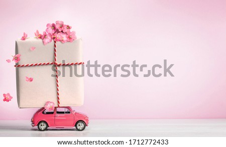 Pink toy retro car with gift box on a roof and flowers on pink background. Copy space.