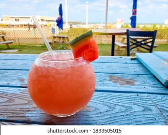 A Pink, Watermelon Based Cocktail In A Fishbowl Glass With A Piece Of Fresh Watermelon. The Drink Is Resting On A Blue, Reclaimed Wood Table By The Ocean.