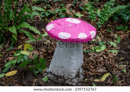 Pink and white painted stone mushroom seat in enchanted woodland fairy garden, surrounded by mature plants, ferns and leaves.