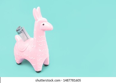 Pink zine type toy alpaca llama with dollar note on blue background close up, coin money bank. Creative and fun trendy collage of funky animal saving for travel concept with copy space